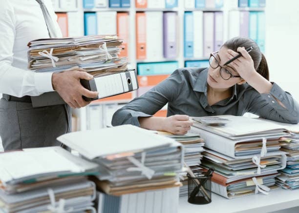 Kanzleiinhaberin zwischen Aktenstapeln, ein Mitarbeiter bringt den nächsten